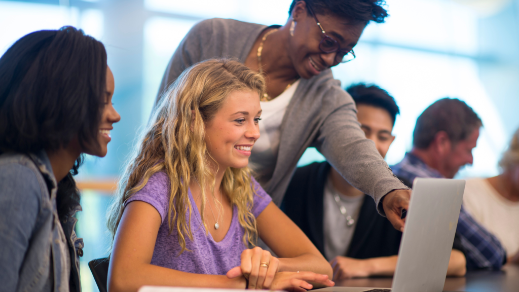 Three students in a row. Professor leans over one and points at her computer screen.