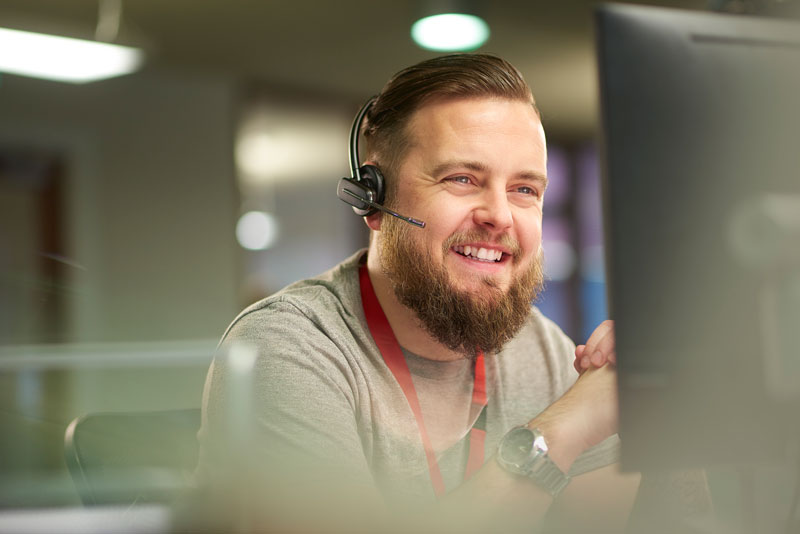 A man in a call center wears a headset and smiles at his computer.